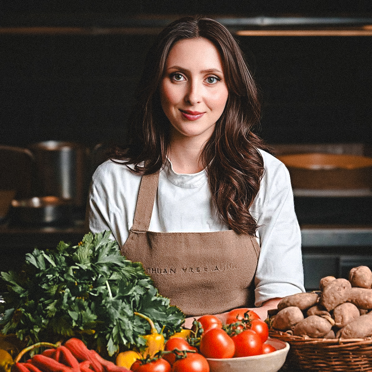 this is an image of taylor in a white chef coat and tan apron standing in a kitchen surrounded by vegetables